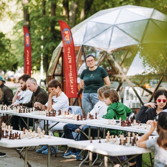 Pro lapus užfiksuotas kadras, kuriame matomos dešimt ant stalų sudėtos šachmatų lentos. Prie kiekvienos iš jų po žmogų, žaidžiantį šachmatų partiją. Dauguma sėdinčių- vaikai. Už žaidėjų stovi moteris ir prižiūri kiekvieną partiją. Prieš vieną žaidėją stov