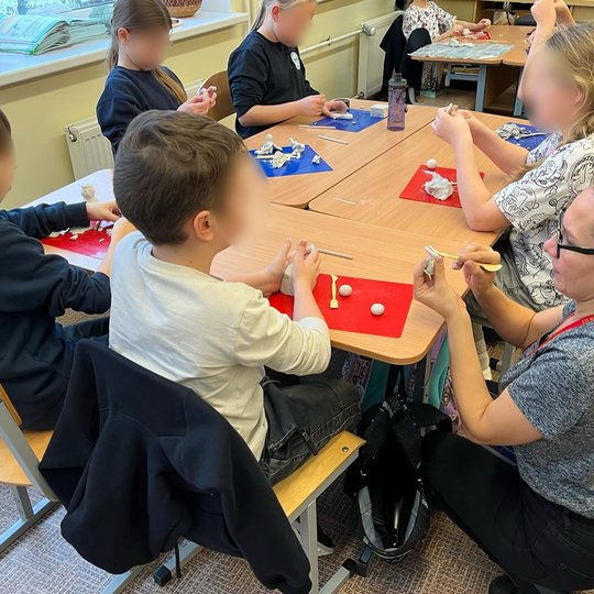 Nuotraukoje prie į didesnį stalą sustumtų trijų mokyklos suolų sėdi ir molį lipdo šeši vaikai. Priešais juos padėtos molio lipdymo lentelės su įvairios formos molio gabalėliais. Prie vieno berniuko pritūpusi moteris su akiniais demonstruoja, kaip iš molio