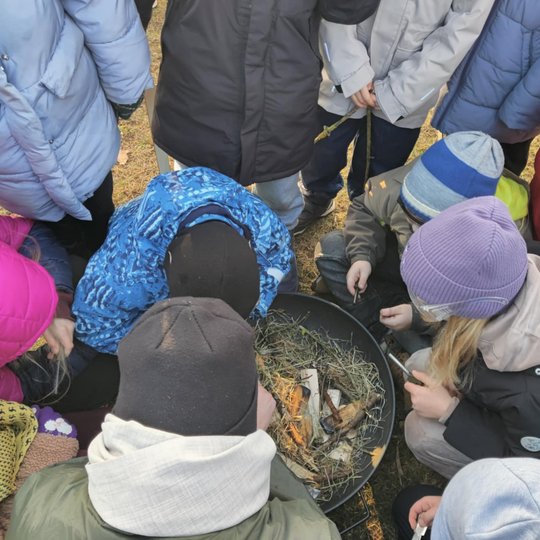 Nuotraukoje matomi vaikai maždaug nuo 7 iki 10 metų, apsirengę šiltai. Penki vaikai yra atsitūpę ir žiūri į puodą prikrauta šieno. Kiti vaikai, kurių matomos tik striukės, stovi šalia.