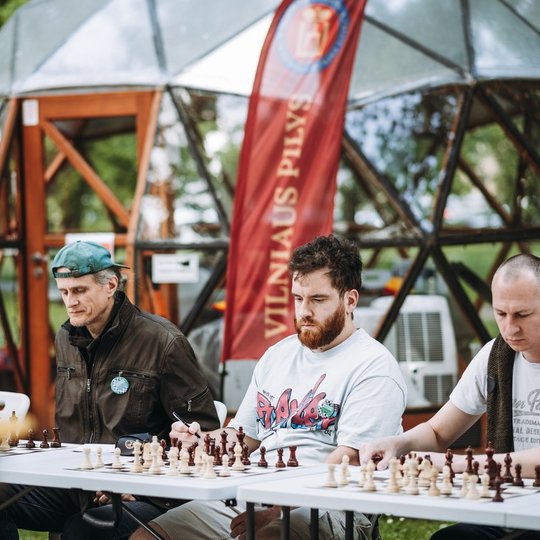 Nuotraukoje matome keturis vyrus ir keturias šachmatų lentas. Vyrai atrodo susimąstę ir labai susikaupę. Už jų fone matome stiklinį kupolą ir vėliavą ant kurios yra parašyta “Vilniaus pilys”. Taip pat kairės pusės apačioje matome išsikišusią moters galvą.