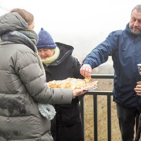 Žmonių grupė lauke dalijasi pyragu – moteris laiko padėklą su kepiniu, vyras ima gabalėlį, kiti rankose laiko popierinius puodelius su gėrimais. Visi šiltai apsirengę žieminiais drabužiais, fone – žiemos peizažas su apniukusiu dangumi.