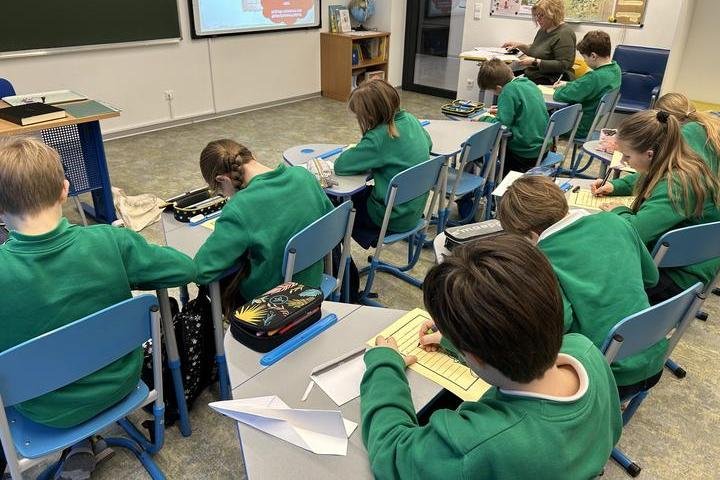 Nuotraukoje vaizduojama klasė. Joje sėdintys vaikai dėvi vienodos spalvos megztinius, visi pasilenkę prie savo stalų kažką rašo. Klasės priekyje yra pakabinta lenta ir išmanusis ekranas ant kurio rodoma prezentacija. Kampe stovi nedidelė lentyna su gaubli