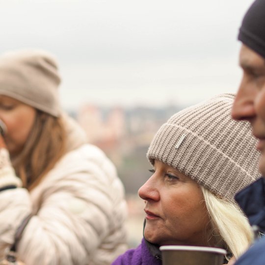 Žmonės su žieminėmis kepurėmis ir pirštinėmis geria šiltus gėrimus lauke. Priekyje – moteris su smėlio spalvos kepure žvelgia į šoną, laikydama popierinį puodelį. Fone – kitas žmogus geria iš puodelio, o dar vienas matomas krašte. Atmosfera žiemiška ir ja