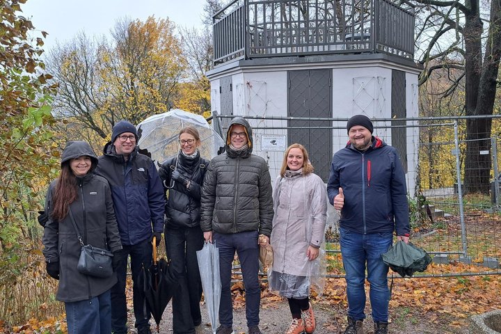 Šeši žmonės, vilkintys rudens drabužiais ir laikantys skėčius, pozuoja prie baltos aštuonkampės Altanos pastato rudeniniame parke.