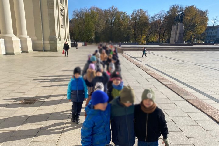 Vaikų grupė žygiuoja išsirikiavusi eile šalia didingų kolonų aikštėje, fone matyti Gedimino paminklas ir medžiai su rudeniniais lapais