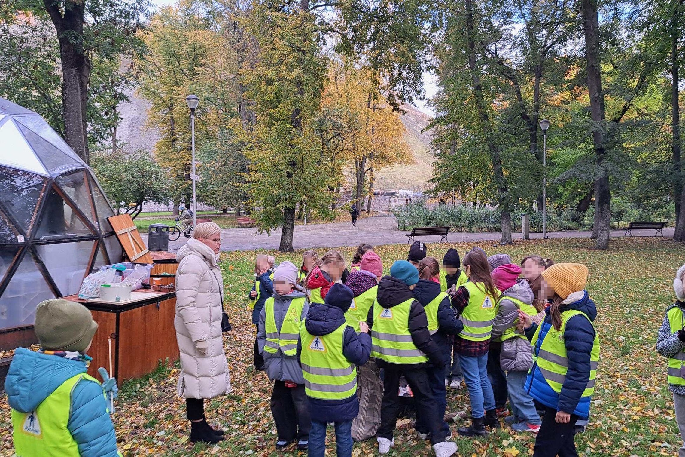 Vaikų grupė su suaugusia lydinčiąja asmenimi stovi parko teritorijoje šalia stiklinio kupolo, rudeniniai lapai nukloję žemę.