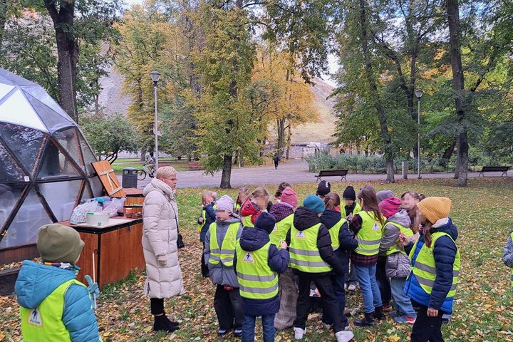 Vaikų grupė su suaugusia lydinčiąja asmenimi stovi parko teritorijoje šalia stiklinio kupolo, rudeniniai lapai nukloję žemę.