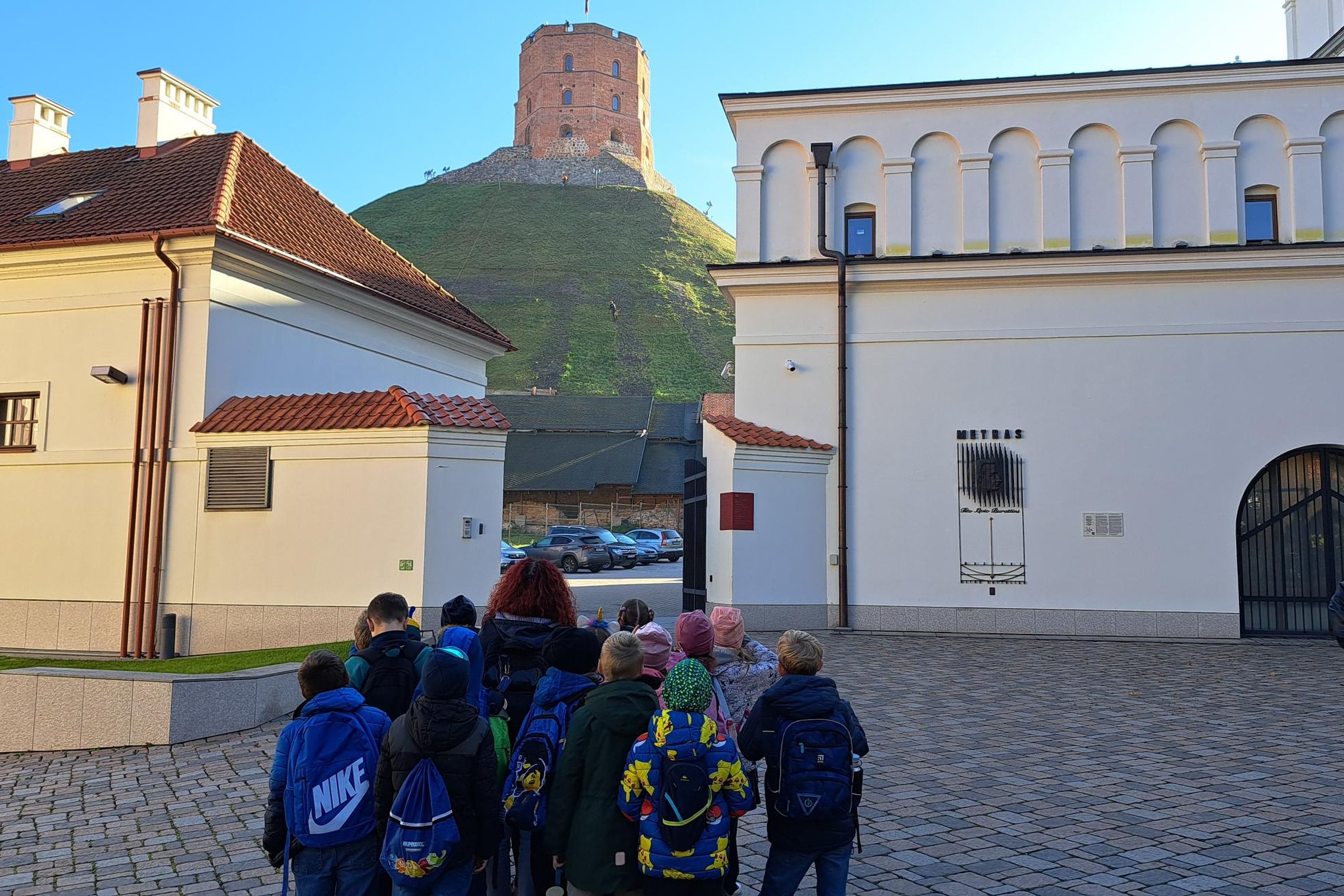 Vaikų grupė su kuprinėmis stovi Vilniaus pilių teritorijoje, fone matomas Gedimino bokštas ant kalno ir balti istoriniai pastatai.