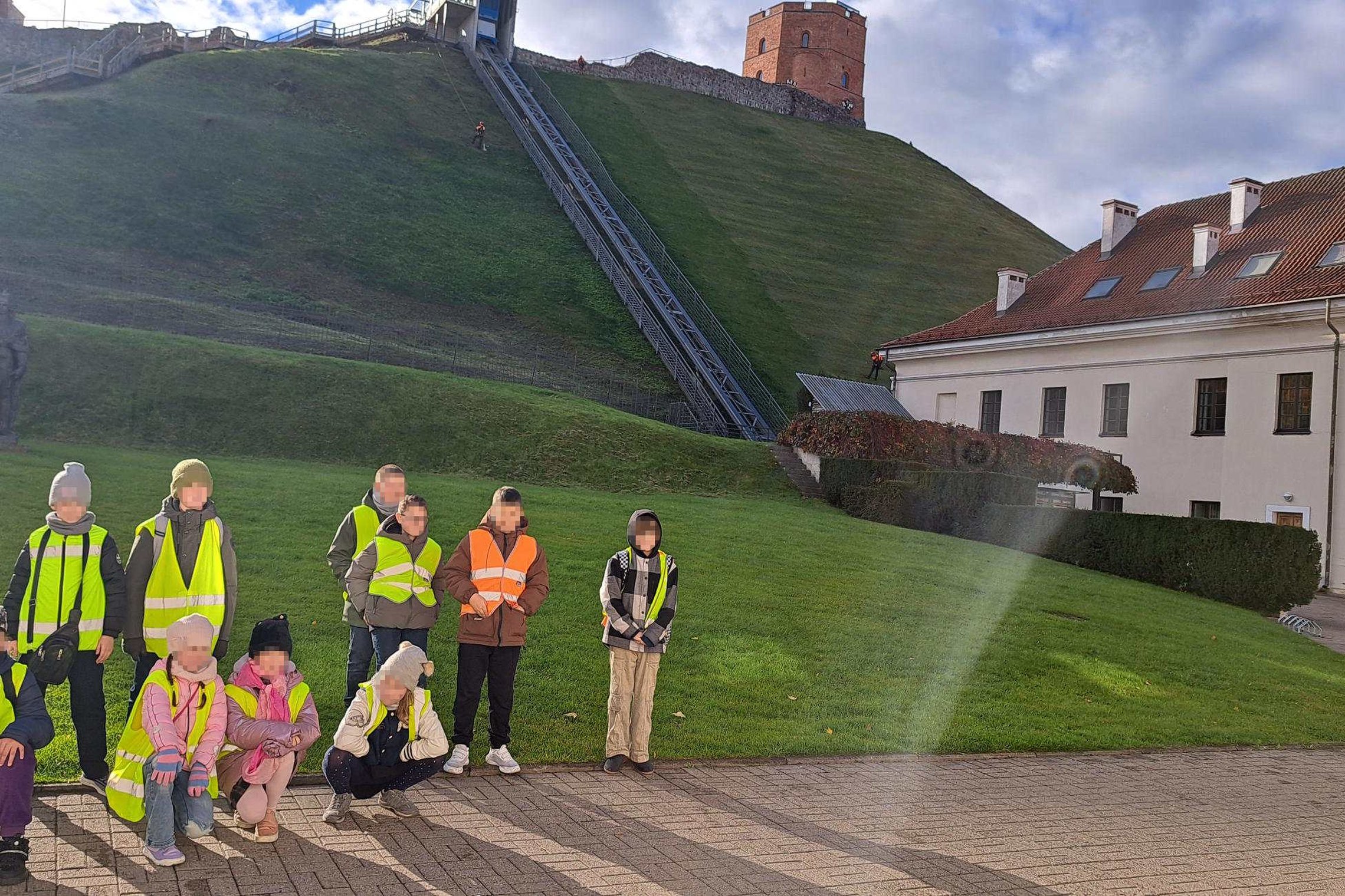 Vaikų grupė su saugos liemenėmis pozuoja priešais Gedimino kalną, funikulierių ir pilies bokštą saulėtą dieną.