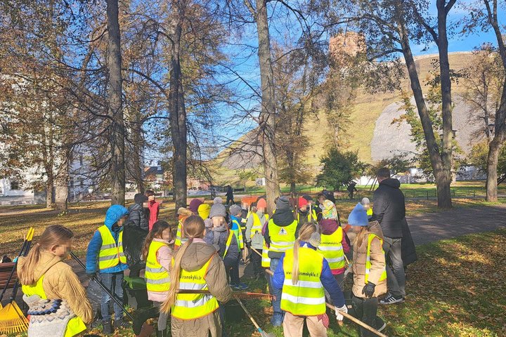Vilniaus šiuolaikinio mokyklos centro trečios klasės mokiniai su atšvaitinėmis liemenėmis dalyvauja edukacijoje prie Gedimino kalno, grėbia rudeninius lapus ir kuria savo piliakalnius, fone matyti Gedimino pilies bokštas.