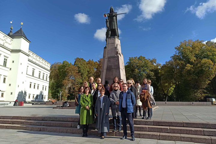 Nuotraukoje grupė žmonių dalyvauja ekskursijoje, stovi ir klausosi gido prie paminklo valdovui Gediminui.