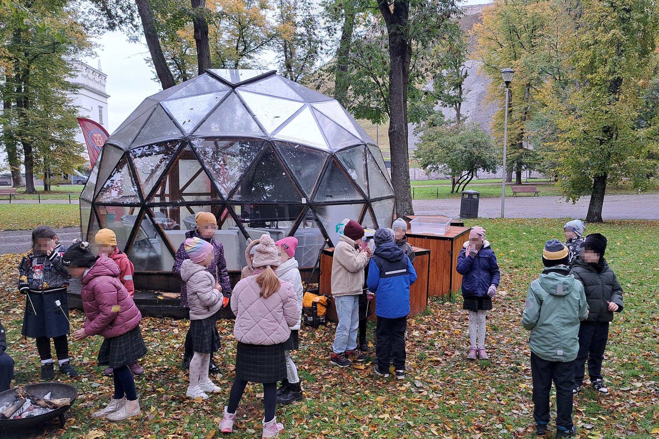 Vaikai su mokytoja rudeniniame parke prie stiklinio kupolo, šalia ugnies dubenų edukacijos metu apie ugnies kelią per amžius.