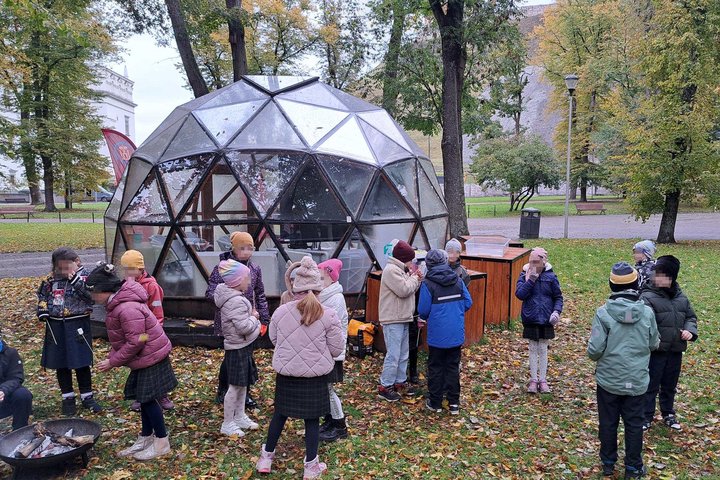 Vaikai su mokytoja rudeniniame parke prie stiklinio kupolo, šalia ugnies dubenų edukacijos metu apie ugnies kelią per amžius.