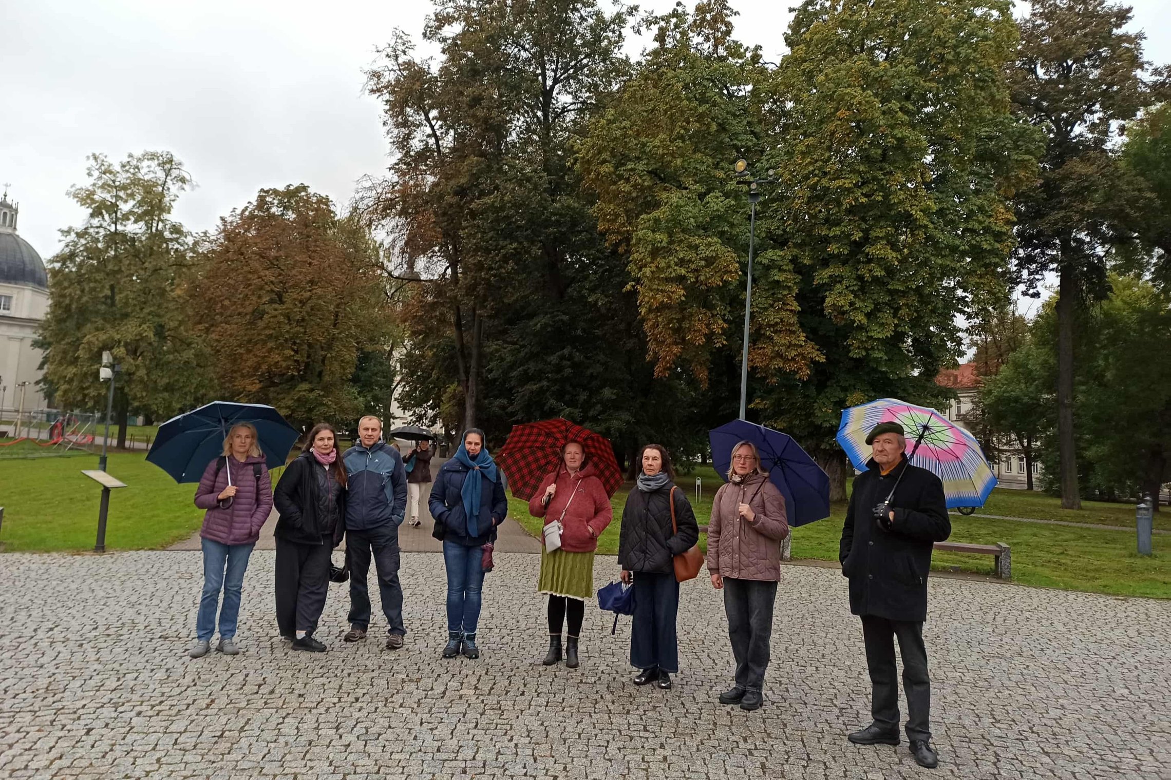 Nuotraukoje nedidelė žmonių grupė dalyvauja ekskursijoje. Visi su skėčiais ir šiltai apsirengę, nes šalta.