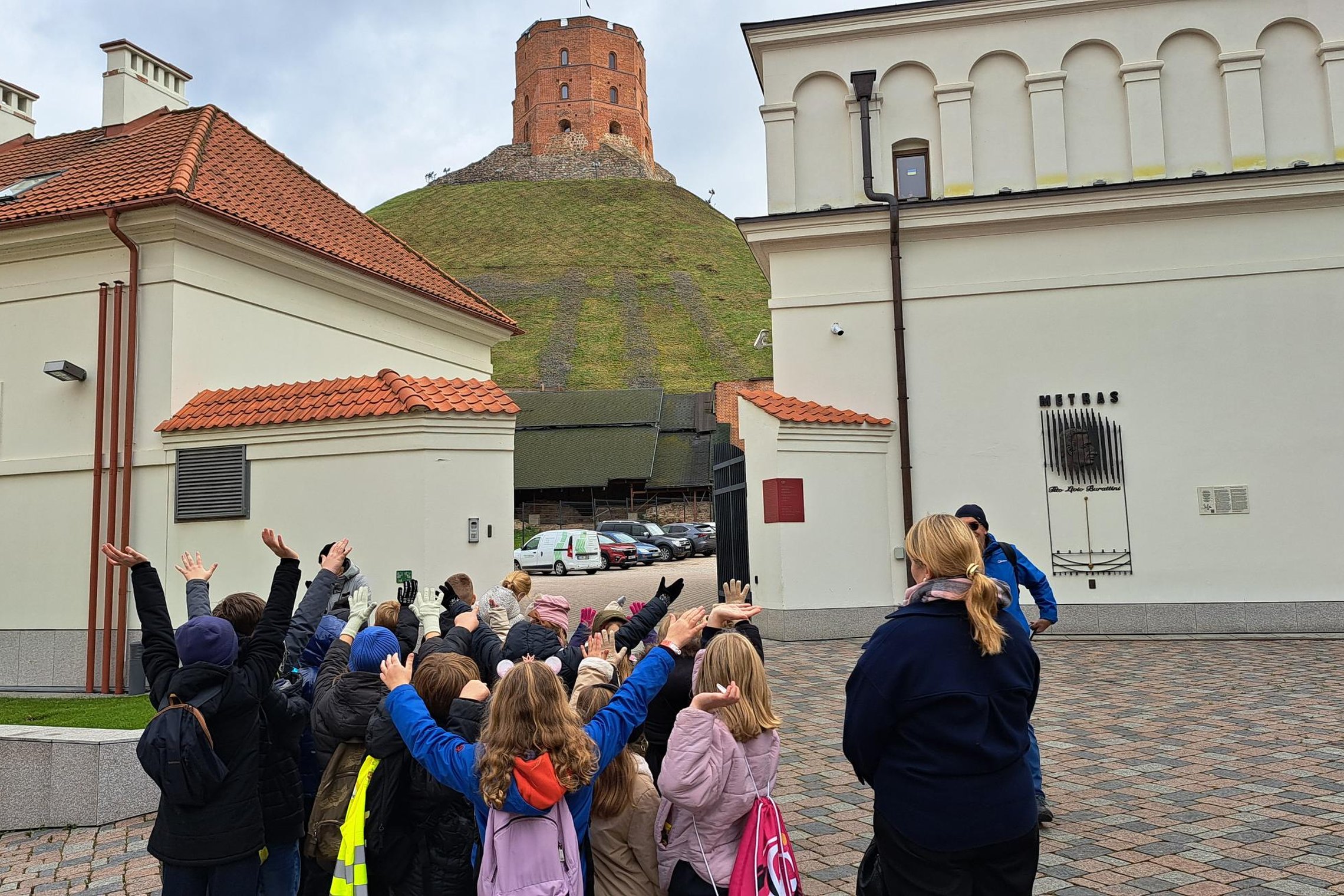 Vaikų grupė su mokytoja kieme prie Vilniaus gynybinės sienos, vaikai pakėlę rankas į Gedimino pilies bokštą kalno viršuje.
