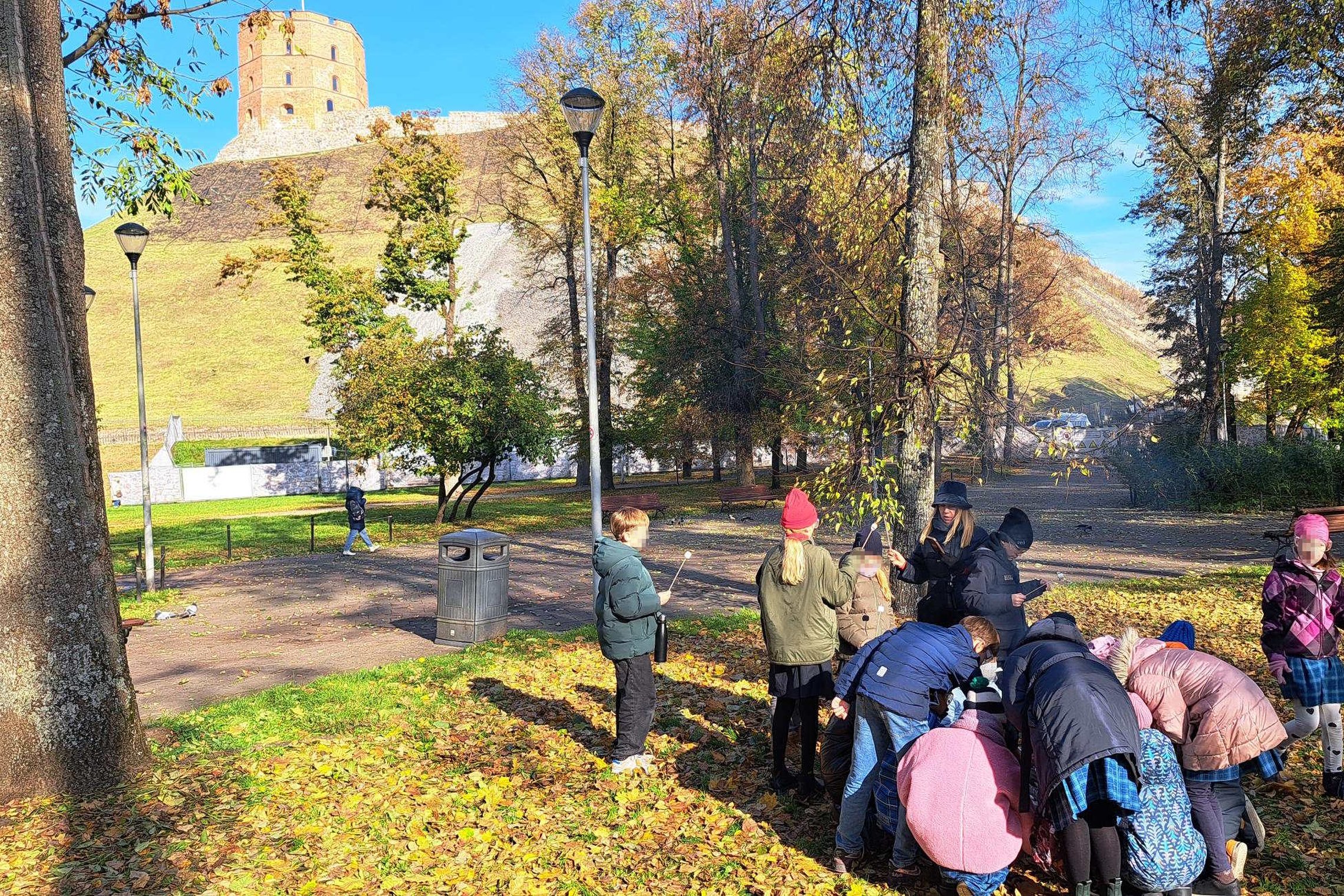 Vaikų grupė su mokytoja dalyvauja edukacijoje „Nuo išlikimo iki žinios: ugnies kelias per amžius“ prie Gedimino kalno papėdės, rudeninėje aplinkoje ant žemės matyti lapų danga ir fone iškilęs Gedimino pilies bokštas.