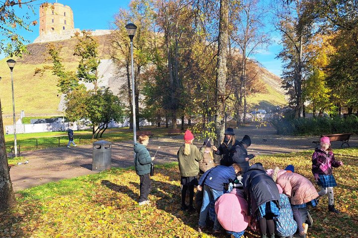 Vaikų grupė su mokytoja dalyvauja edukacijoje „Nuo išlikimo iki žinios: ugnies kelias per amžius“ prie Gedimino kalno papėdės, rudeninėje aplinkoje ant žemės matyti lapų danga ir fone iškilęs Gedimino pilies bokštas.