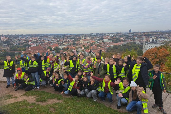Nuotraukoje didelė grupė vaikų mojuoja mokytojoms. Už jų nugarų Vilniaus panorama, kuri atsiveria nuo Plikojo kalno.