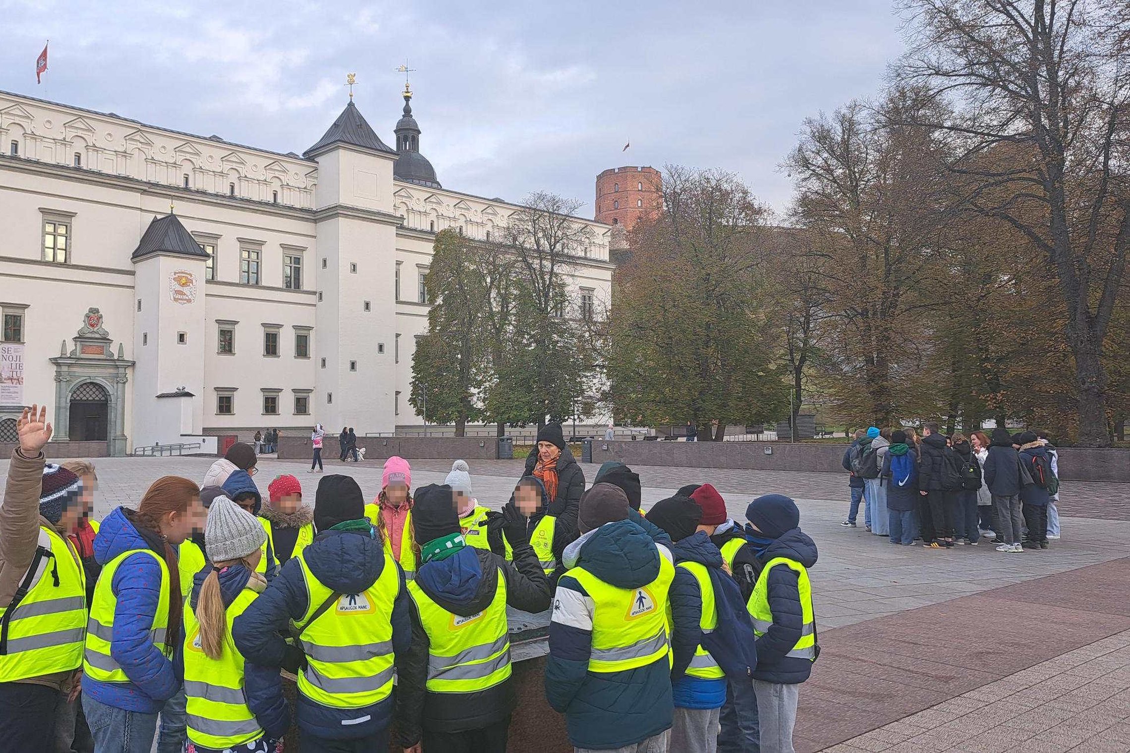 Ketvirtokų grupė su geltonomis atšvaitinėmis liemenėmis dalyvauja edukacijoje „Gediminas ir trys Vilniaus pilys“ prie Valdovų rūmų Vilniuje, fone matyti Gedimino pilies bokštas.