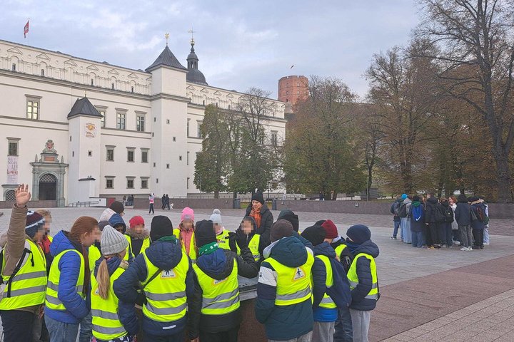 Ketvirtokų grupė su geltonomis atšvaitinėmis liemenėmis dalyvauja edukacijoje „Gediminas ir trys Vilniaus pilys“ prie Valdovų rūmų Vilniuje, fone matyti Gedimino pilies bokštas.