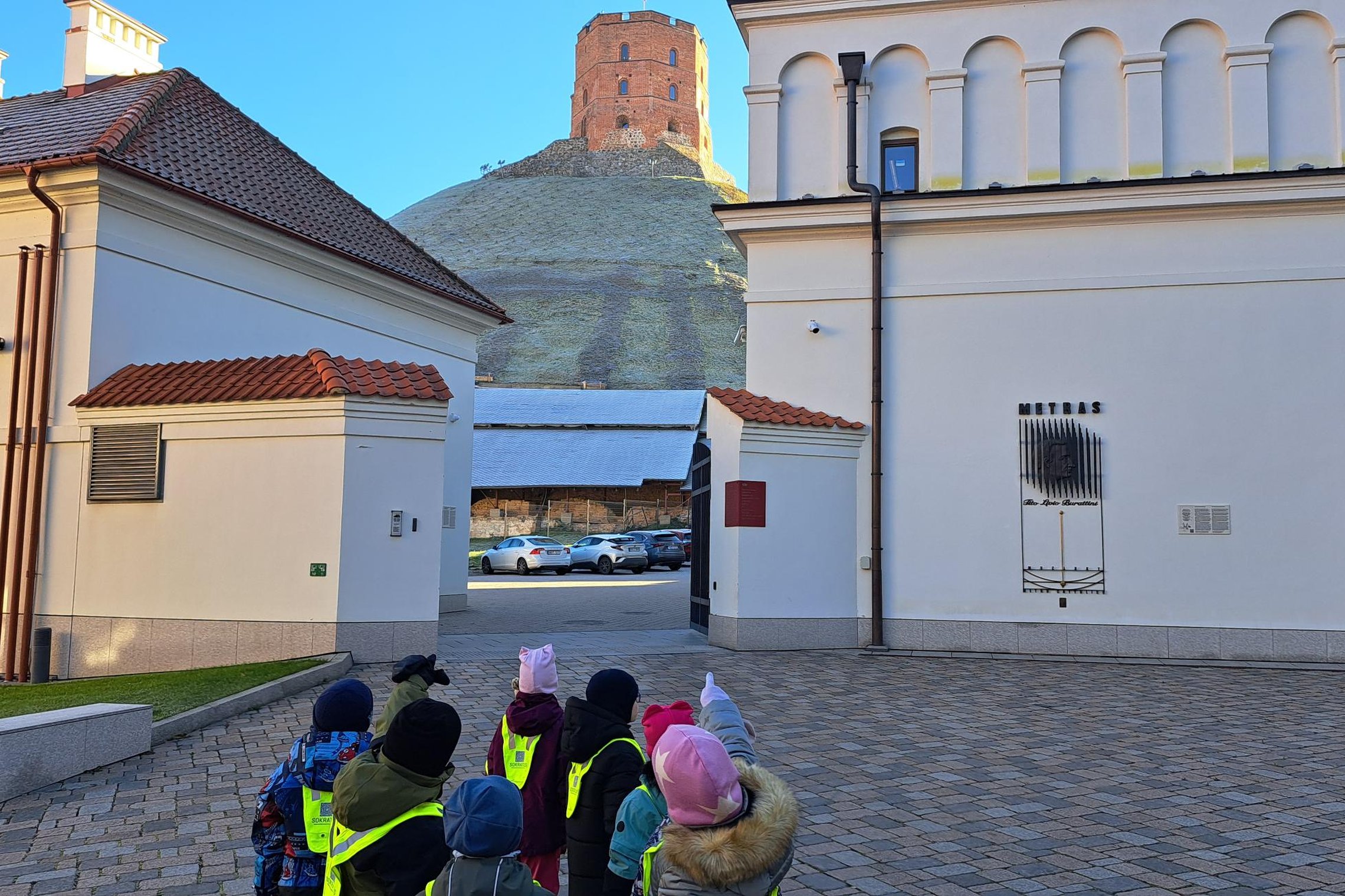 Grupė dešimties vaikų su ryškiais atšvaitais stovi kieme tarp dviejų šviesių pastatų ir rodo į kalną su Gedimino bokštu viršuje.