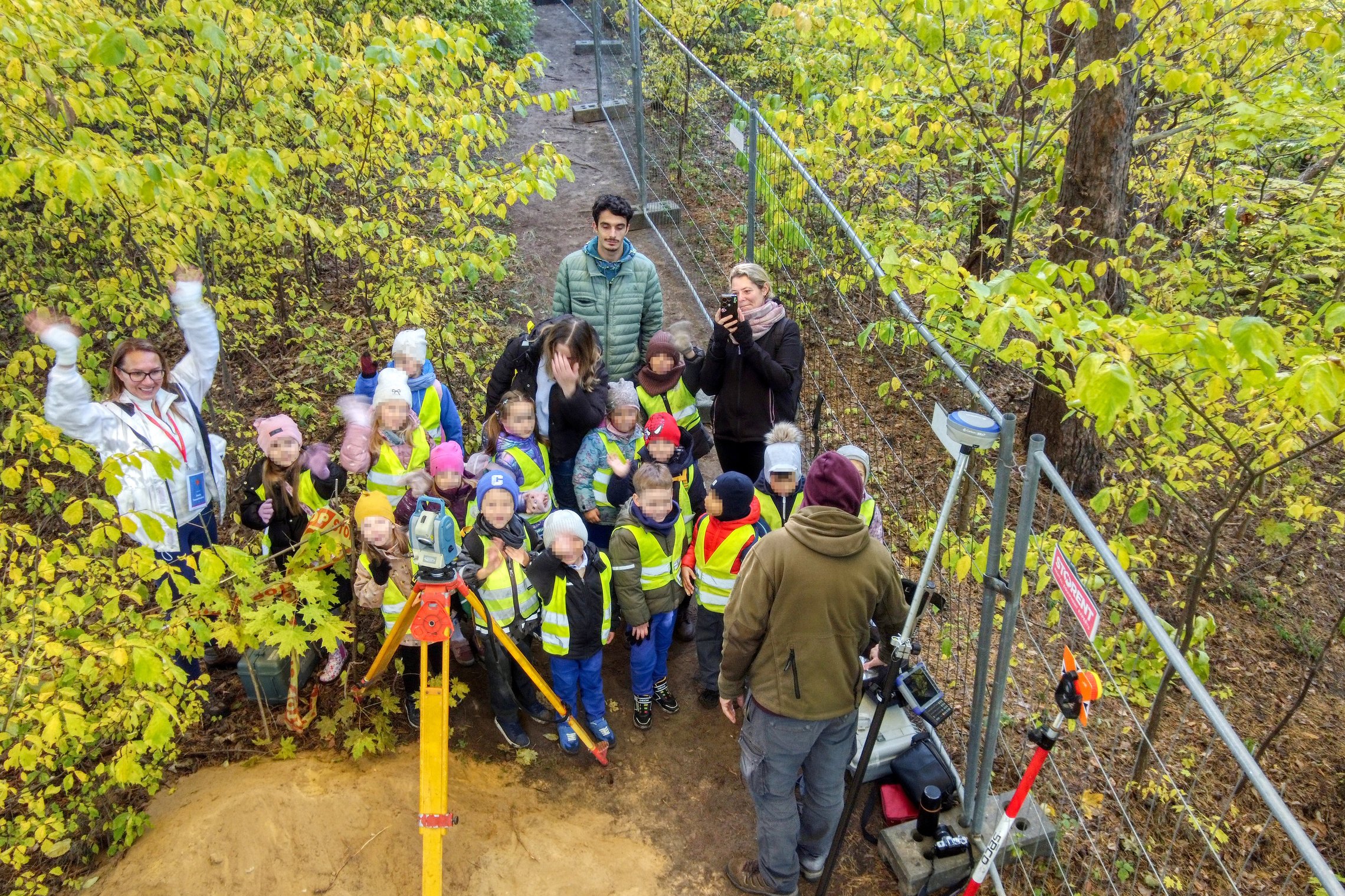 Nuotraukoje vaikų grupė stovi prie archeologinių tyrimų vietos, kurioje dirba archeologai.