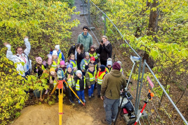 Nuotraukoje vaikų grupė stovi prie archeologinių tyrimų vietos, kurioje dirba archeologai.