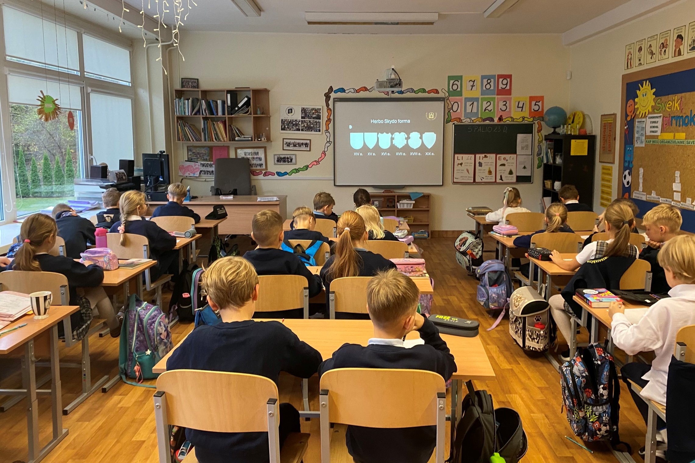 Vaikai klasėje klausosi edukacijos apie heraldiką. Ant ekrano rodoma skaidrė su įvairių laikotarpių herbo skydo formomis. Mokiniai sėdi prie stalų, žiūri į ekraną ir rašo užrašus
