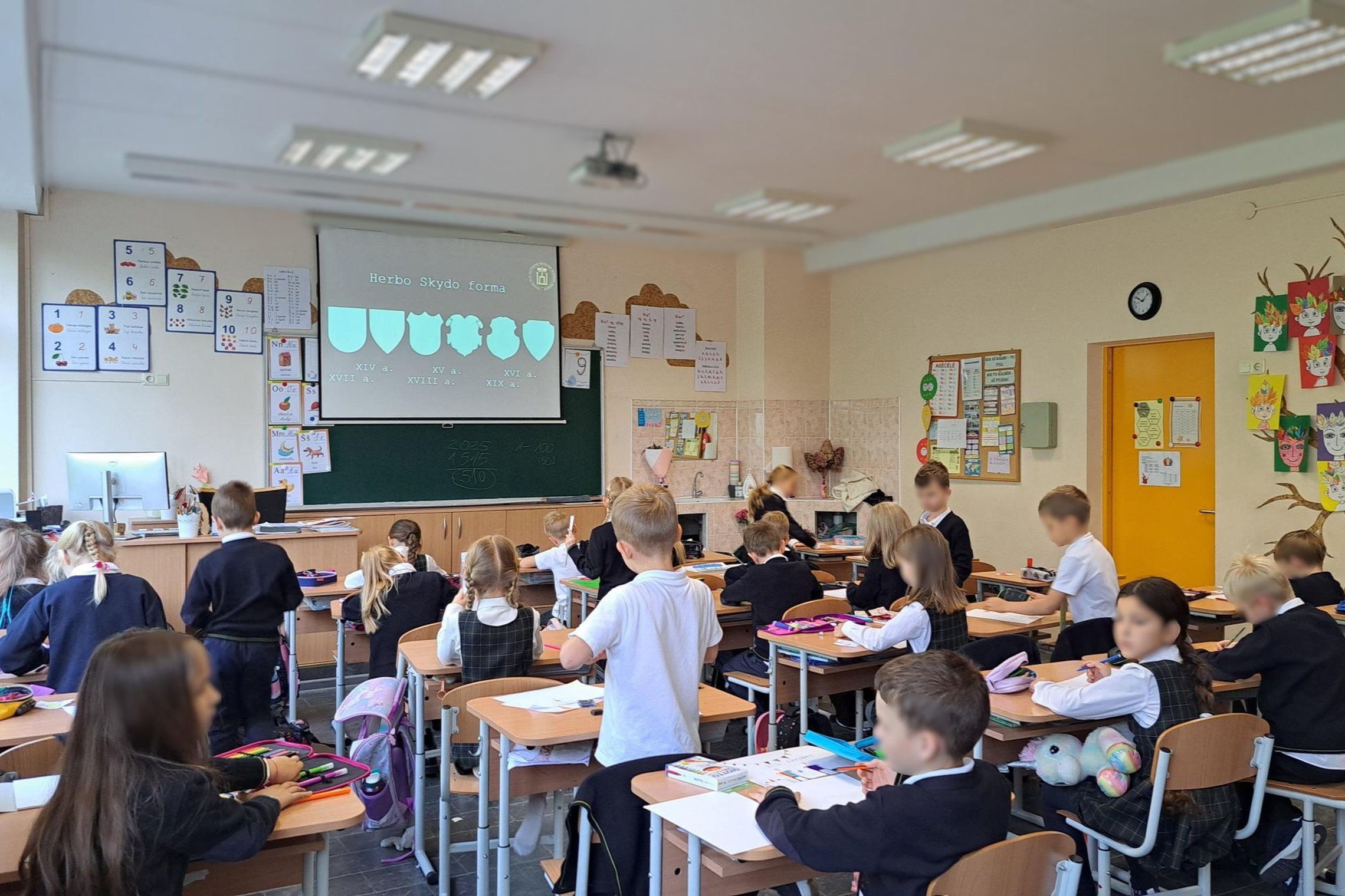 Nuotraukoje vaikai klasėje. Sėdi prie stalų, pasilenkę kuria savo šeimos herbą.
