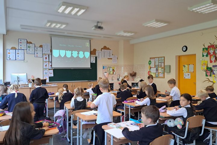 Nuotraukoje vaikai klasėje. Sėdi prie stalų, pasilenkę kuria savo šeimos herbą.
