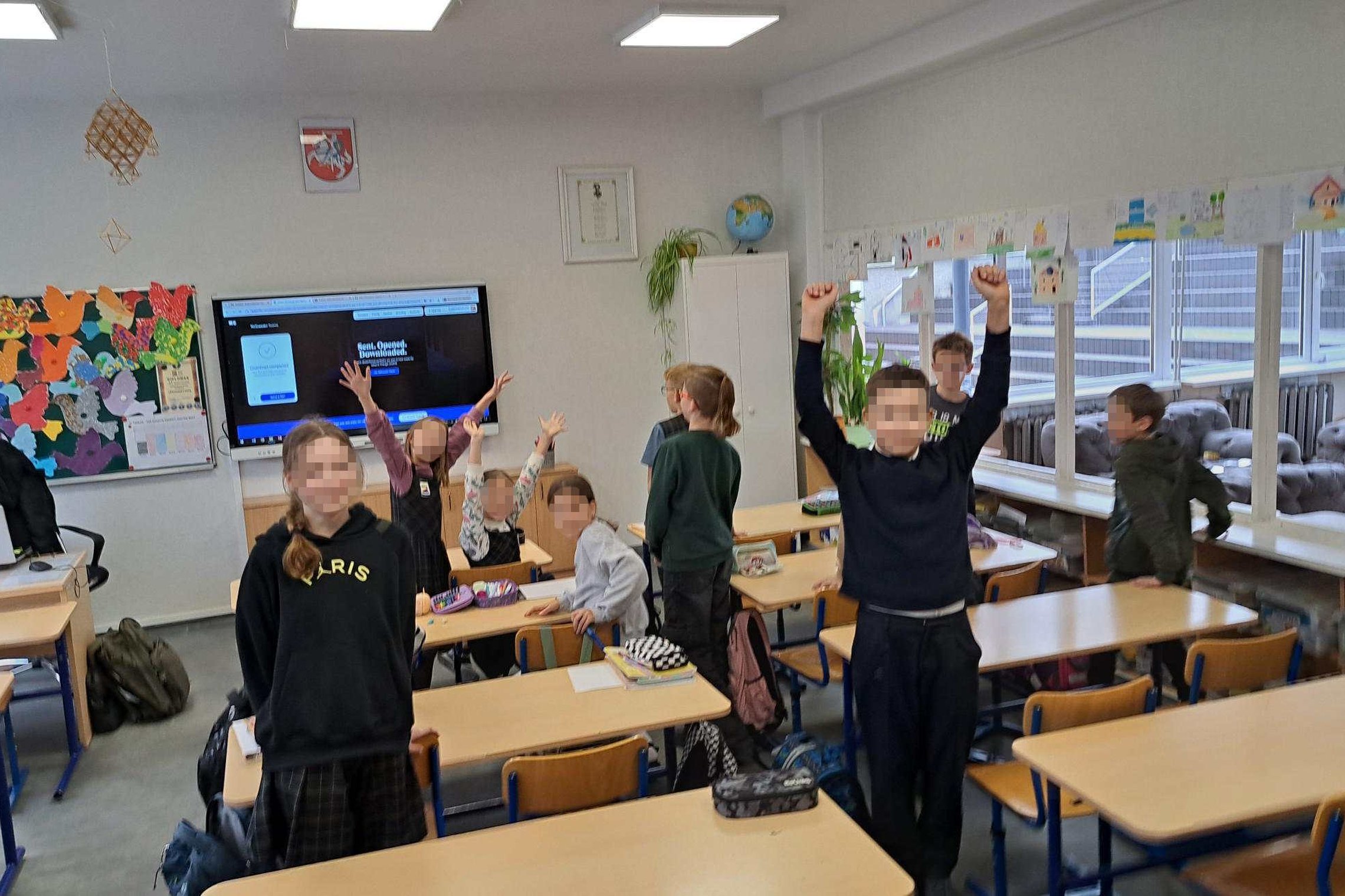 Klasėje vykstančios edukacijos akimirka – keli ketvirtokai stovi prie savo suolų, keli pakėlę rankas į viršų, kiti šypsosi. Fone matyti lenta su spalvingais darbais, ekranas, ant kurio rodomas pristatymas, ir šviesi klasės erdvė su langais.