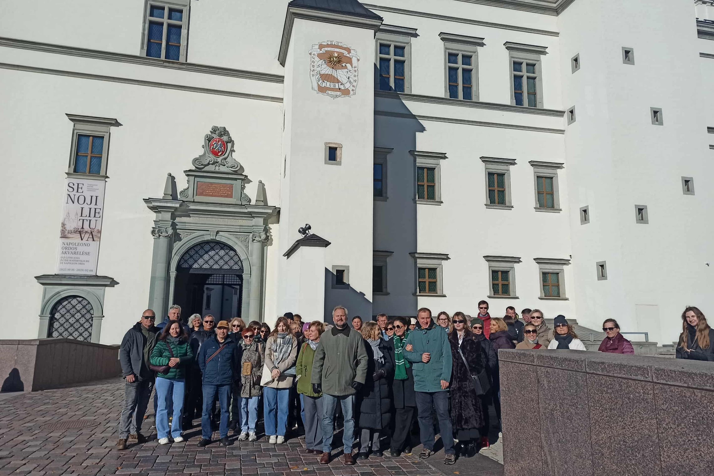 Nuotraukoje didžiulė žmonių grupė dalyvauja ekskursijoje „Vilniaus pilių laikrodžiai“. Už jų nugarų saulės laikrodis.