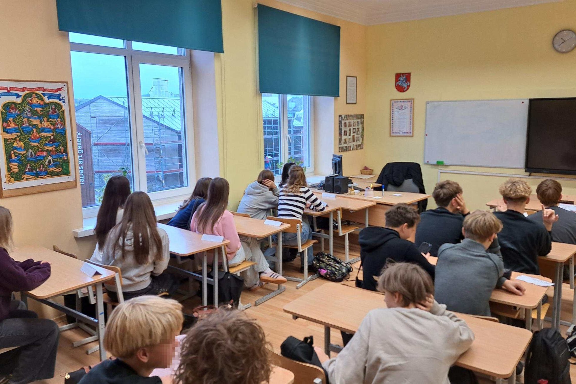 Klasėje prie suolų sėdi devintokai, klausantys edukacijos apie baltų tikėjimą; ant sienos kabo genealoginis medžio tipo plakatas, už langų matosi Užupio pastatai.