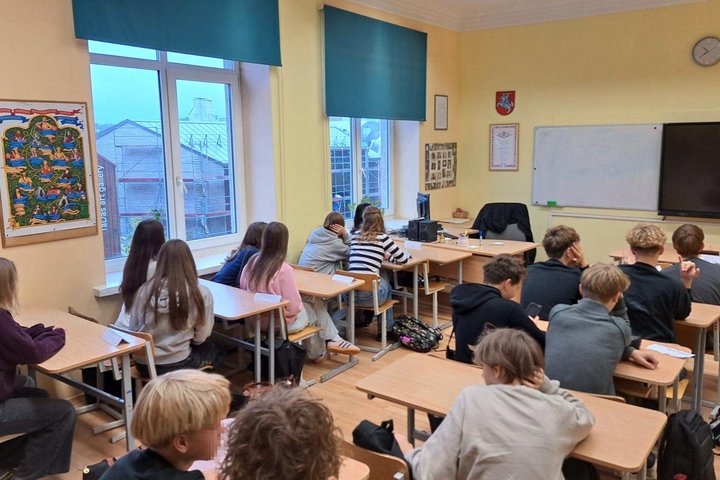 Klasėje prie suolų sėdi devintokai, klausantys edukacijos apie baltų tikėjimą; ant sienos kabo genealoginis medžio tipo plakatas, už langų matosi Užupio pastatai.