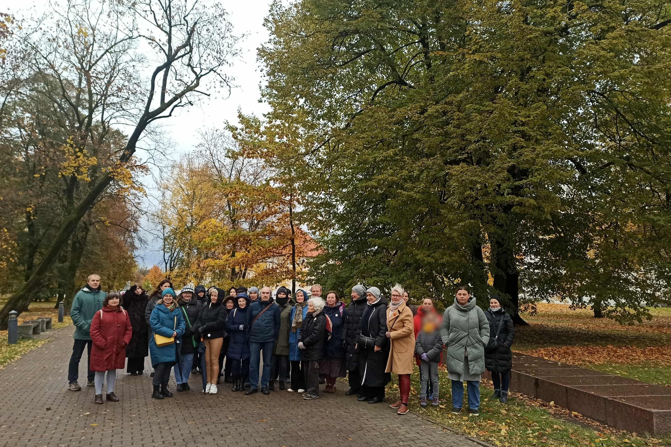 Grupė žmonių, sustojusių parke rudenį, apsuptų spalvotų medžių ir nukritusių lapų. Jie šypsosi ir žvelgia į kamerą, fone matyti takas, vedantis tolyn tarp medžių