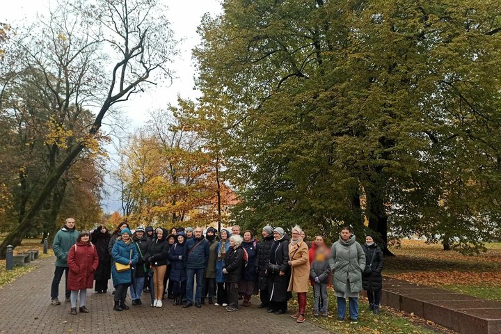 Grupė žmonių, sustojusių parke rudenį, apsuptų spalvotų medžių ir nukritusių lapų. Jie šypsosi ir žvelgia į kamerą, fone matyti takas, vedantis tolyn tarp medžių