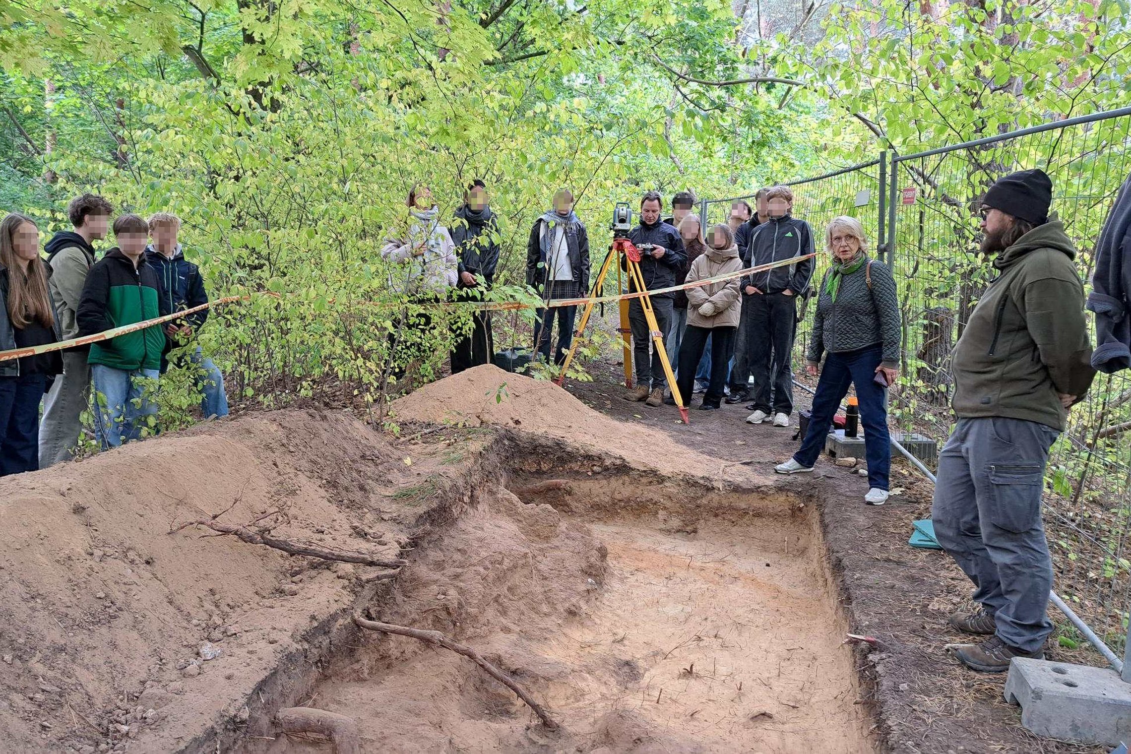 Žmonių grupė stebi archeologinius kasinėjimus miške, šalia iškastos žemės duobės stovi archeologas ir aiškina radinių paieškos procesą.