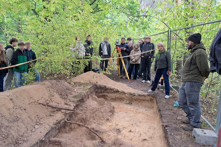 Žmonių grupė stebi archeologinius kasinėjimus miške, šalia iškastos žemės duobės stovi archeologas ir aiškina radinių paieškos procesą.