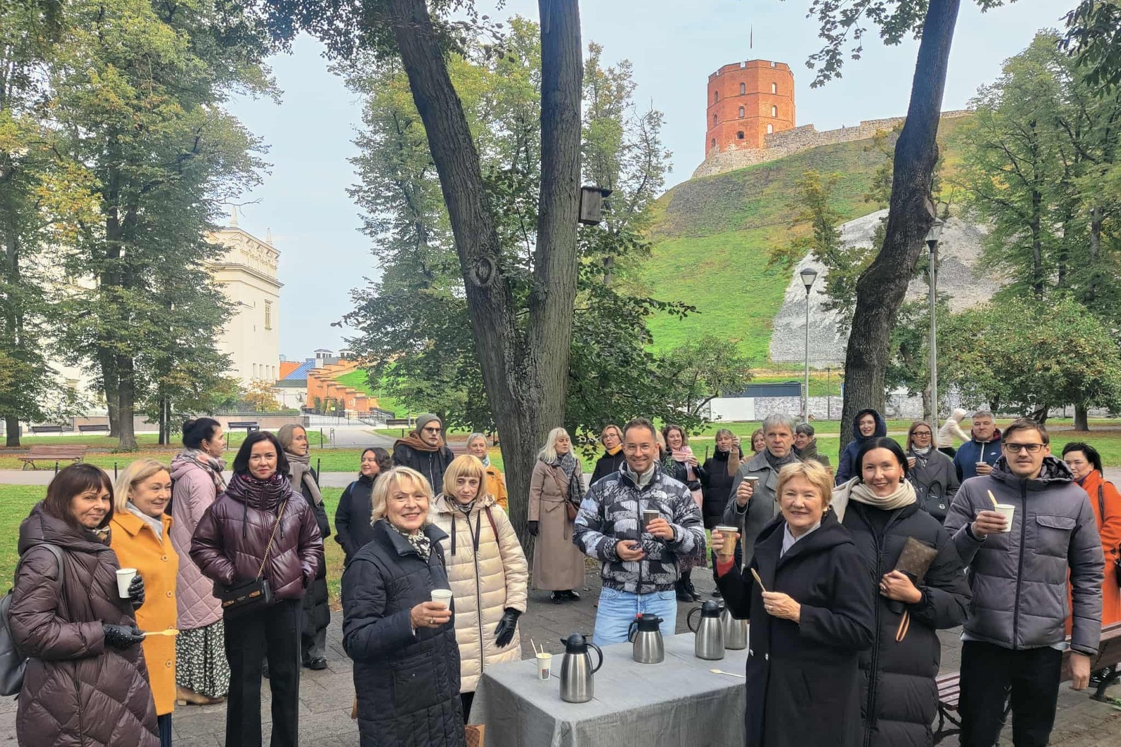 Žmonių grupė lauke prie Gedimino kalno Vilniuje, su matomu Gedimino bokštu fone. Dalyviai stovi prie stalelio su termosais, laiko popierinius puodelius su gėrimais ir šypsosi kamerai. Aplink medžiai ir žalias kalno šlaitas.