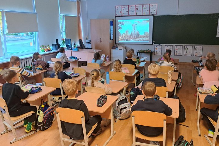 Pradinės klasės mokiniai sėdi suoluose ir žiūri į ekrane rodomą Vilniaus pilių iliustraciją – vyksta edukacija apie Gedimino sapną ir Vilniaus pilis.