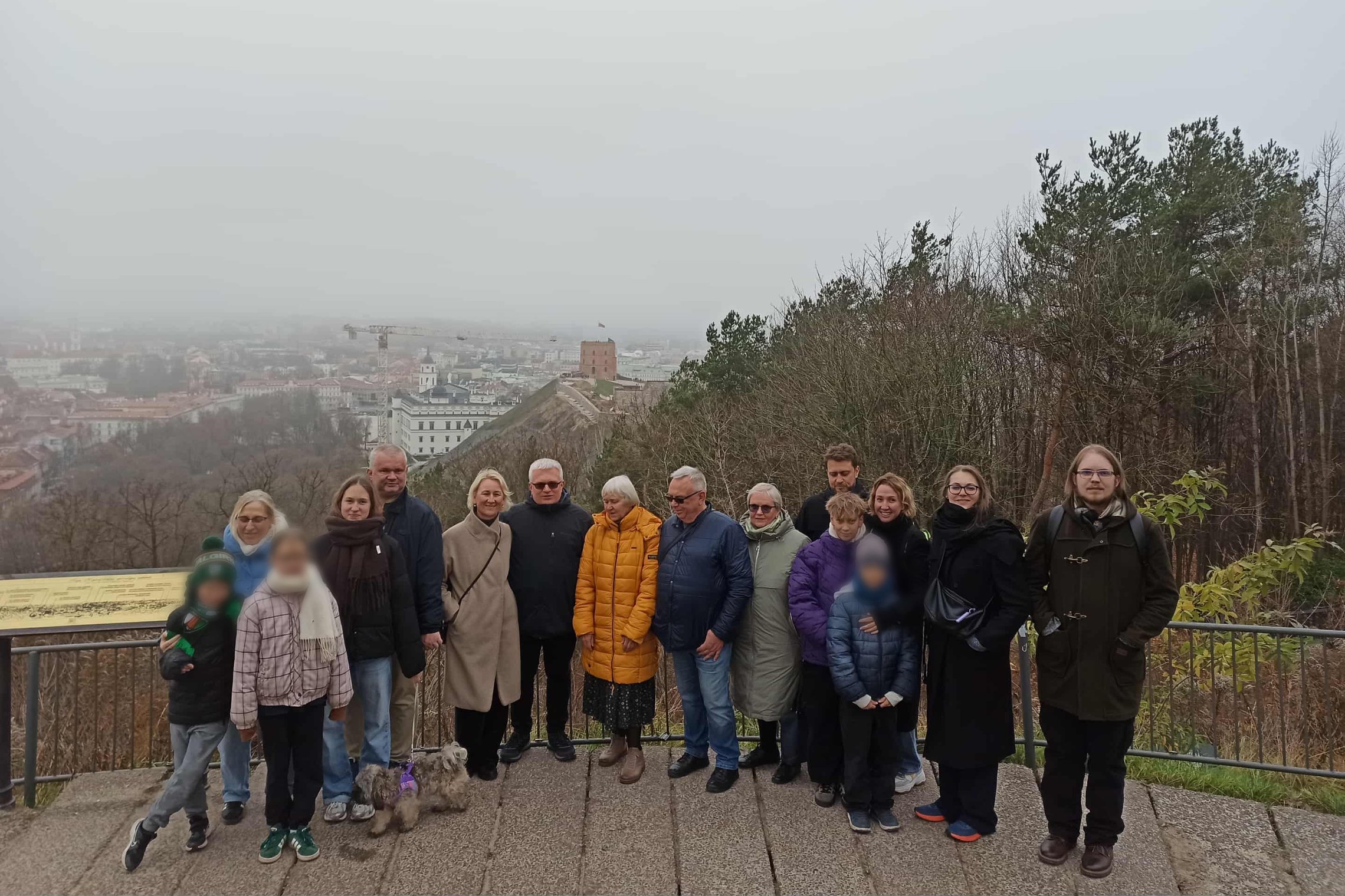 Žmonių grupė stovi apžvalgos aikštelėje virš miesto, fone matyti apsiniaukęs Vilniaus peizažas ir Gedimino bokštas. Priekyje keli vaikai ir šuo, visi apsirengę šiltais drabužiais
