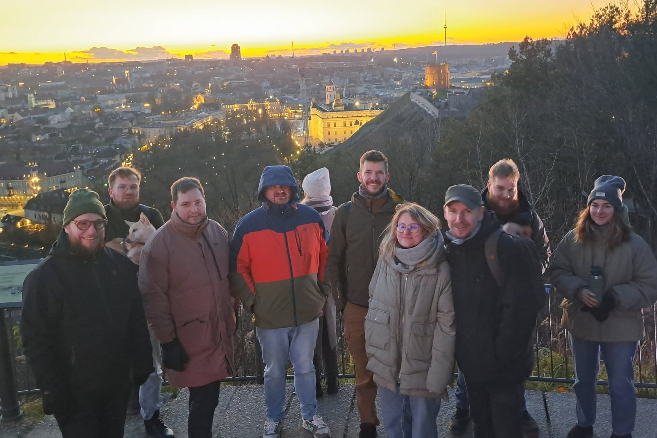 Grupė žmonių stovi ant apžvalgos aikštelės, už jų matyti saulėlydžio nušviesta Vilniaus panorama su Gedimino bokštu ir miesto pastatais.