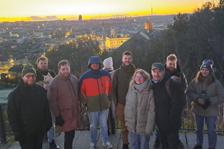 Grupė žmonių stovi ant apžvalgos aikštelės, už jų matyti saulėlydžio nušviesta Vilniaus panorama su Gedimino bokštu ir miesto pastatais.
