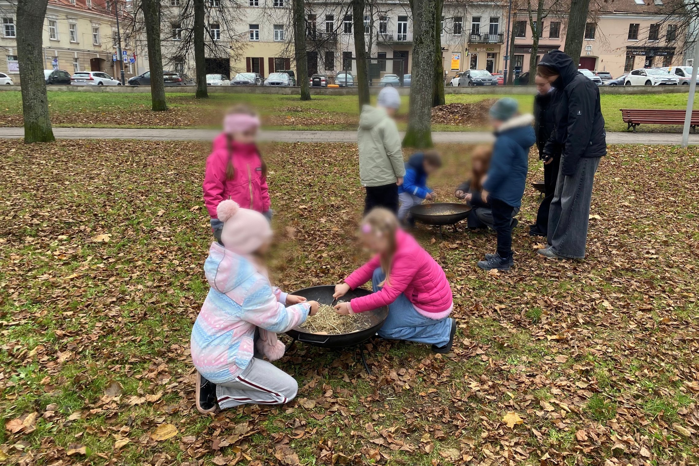 Vaikai lauke, apsupti rudens lapų, grupelėmis mokosi įžiebti ugnį, dirbdami prie metalinių ugniakurų su šiaudais ir pagaliukais