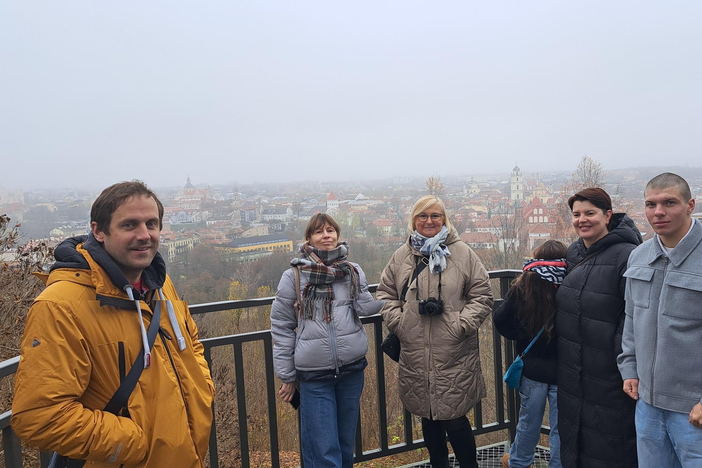 Grupė žmonių stovi Altonos bokštelio apžvalgos aikštelėje, už jų atsiveria rūke paskendęs Vilniaus senamiesčio vaizdas.