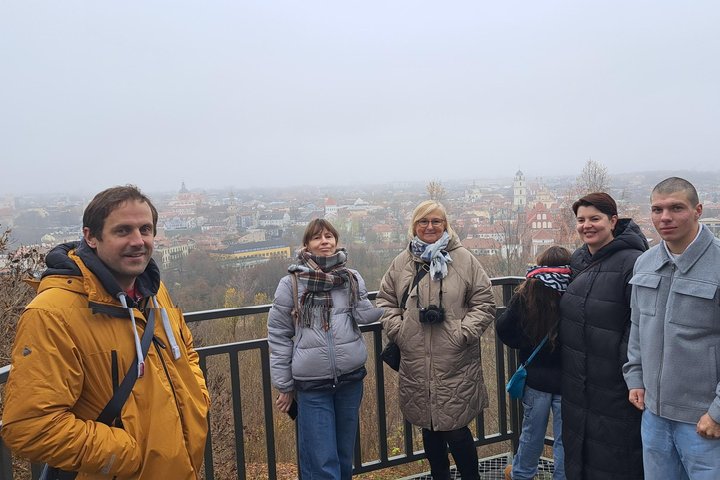 Grupė žmonių stovi Altonos bokštelio apžvalgos aikštelėje, už jų atsiveria rūke paskendęs Vilniaus senamiesčio vaizdas.