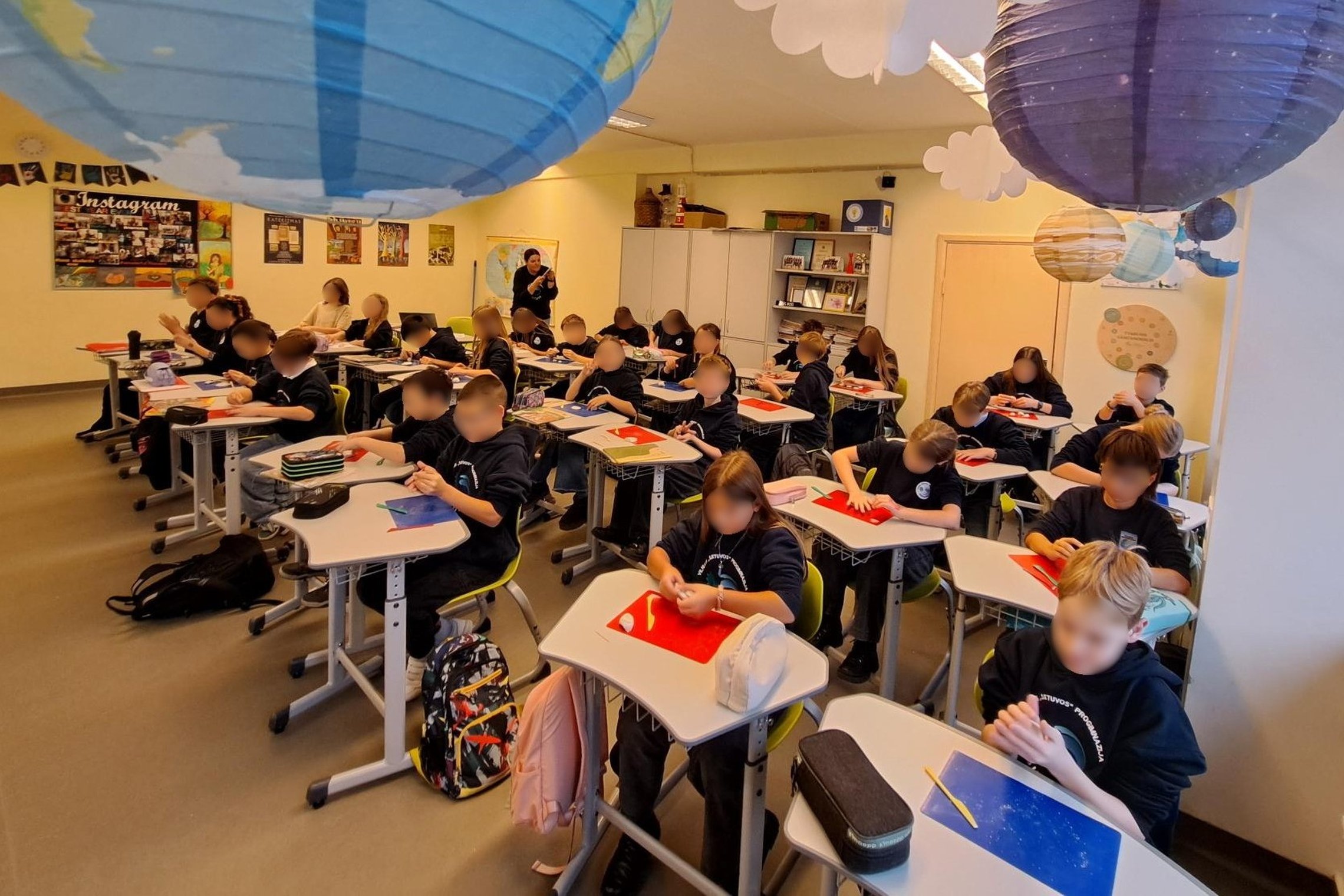 Nuotraukoje klasė, pilna suolų ir vaikų. Panašu, kad istorijos kabinetas. Vaikai sėdi suoluose ir kažką pasilenkia dirba.