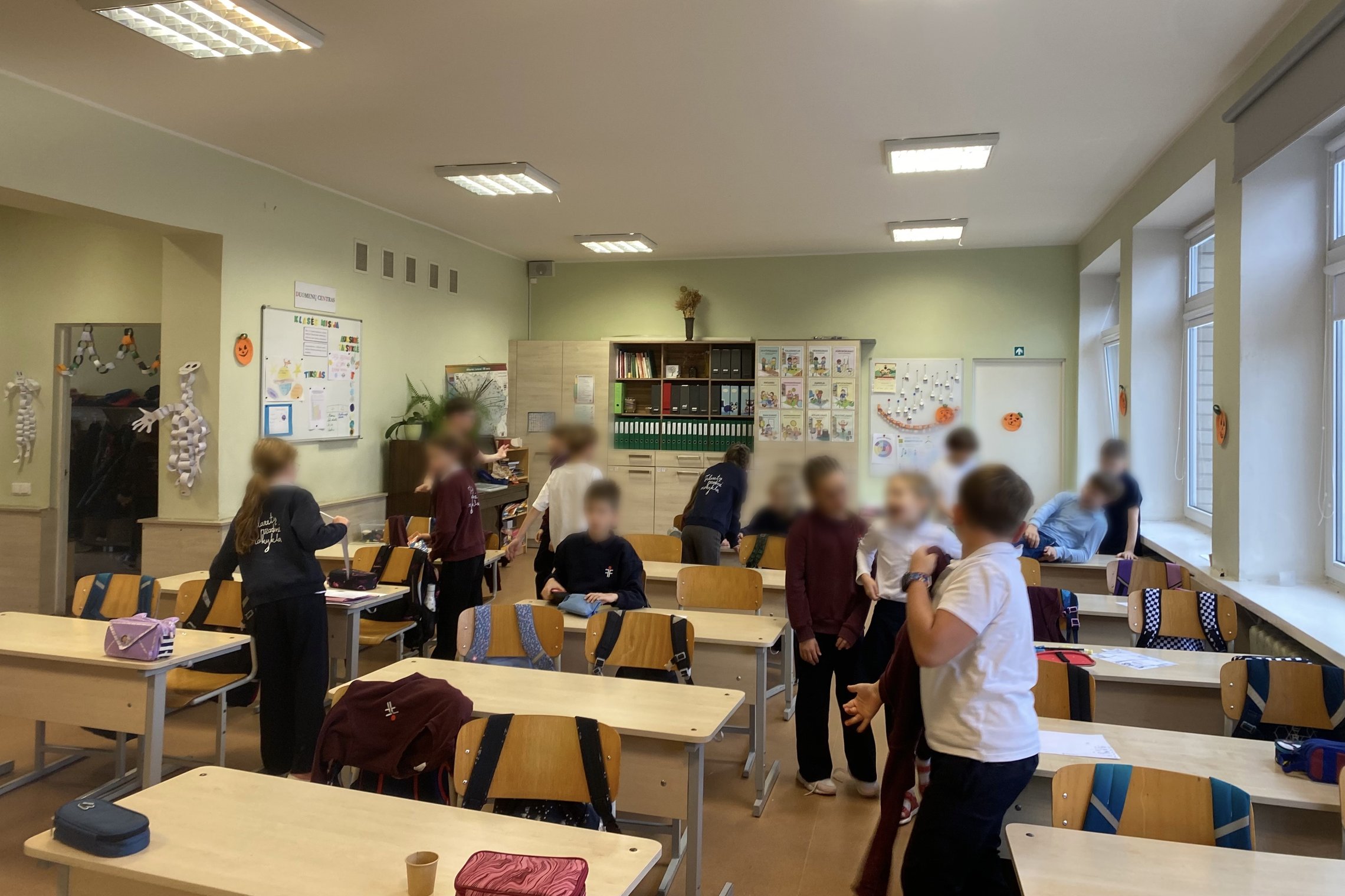 Klasės vaizdas, kuriame grupė vaikų juda ir bendrauja tarp suolų šviesioje mokyklos klasėje. Ant sienų matyti vaikų darbai ir dekoracijos, o ant stalų – mokykliniai reikmenys