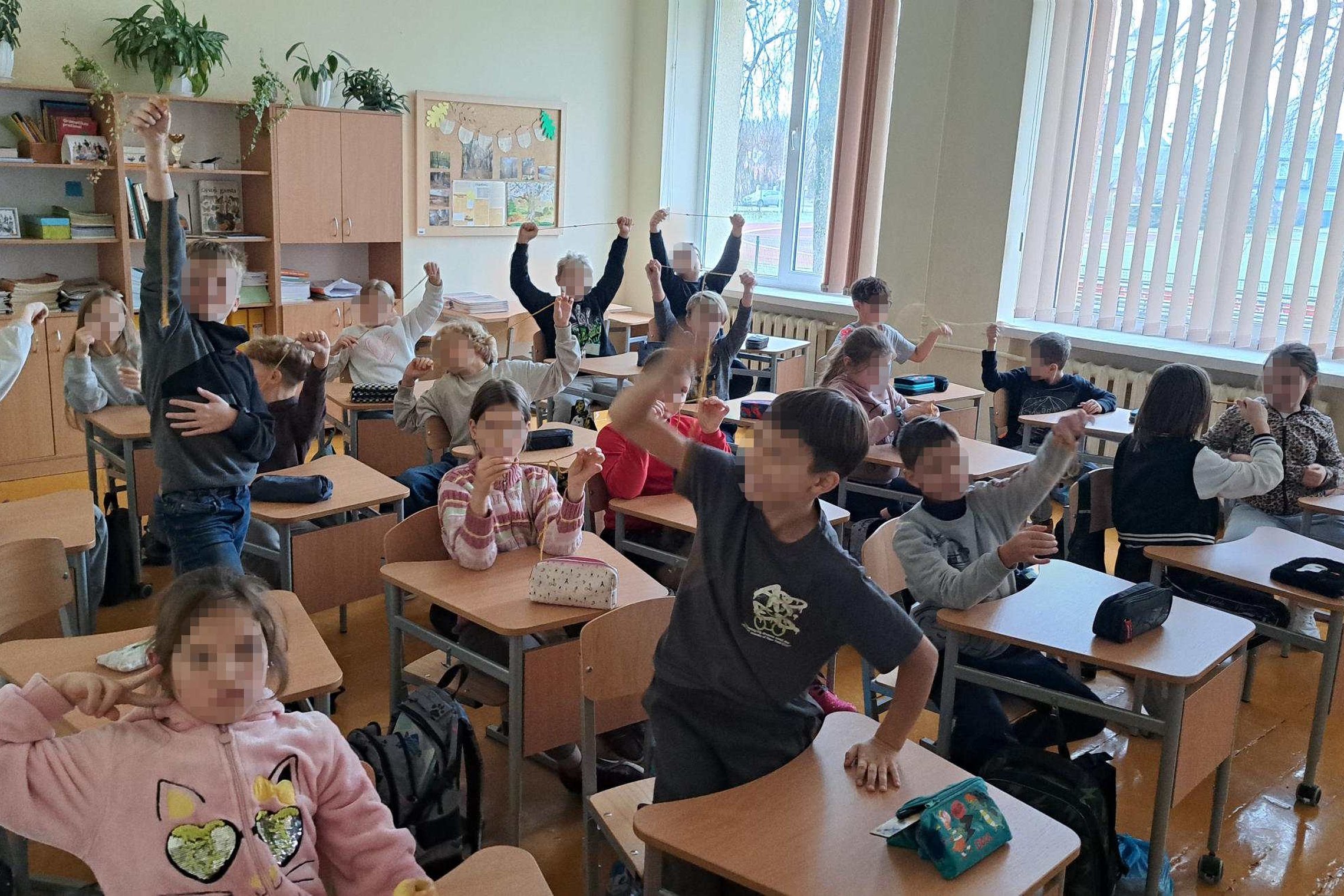 Klasėje sėdintys pradinukai su pakeltomis rankomis riša virveles ir demonstruoja mazgus; aplink matyti suolai, kuprinės, spintos su knygomis ir dideli langai.