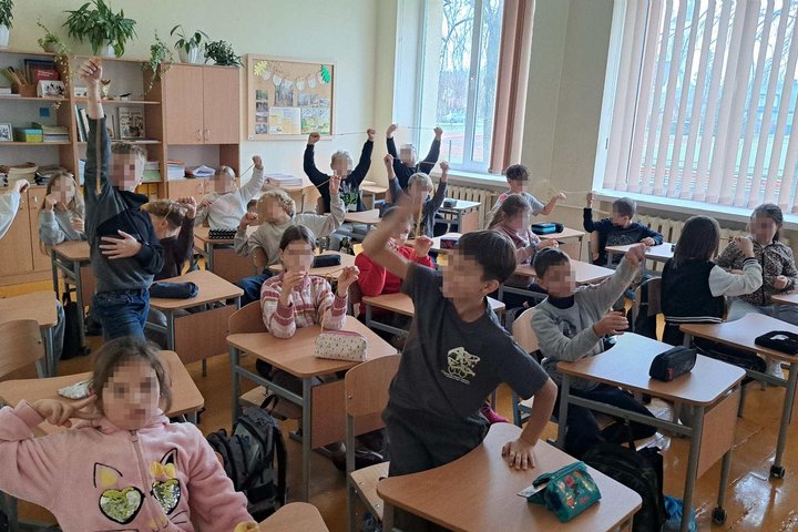Klasėje sėdintys pradinukai su pakeltomis rankomis riša virveles ir demonstruoja mazgus; aplink matyti suolai, kuprinės, spintos su knygomis ir dideli langai.